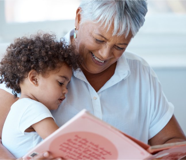 Older woman wearing a white shirt holding a young child while reading a large pink book indoors.
