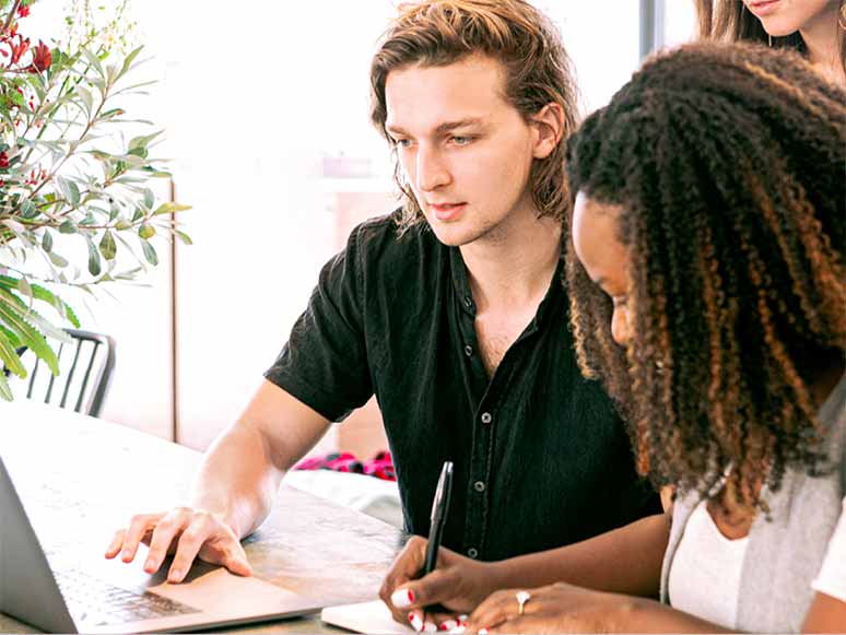 Man and woman collaborating at a table, reviewing something on a laptop while the woman takes notes