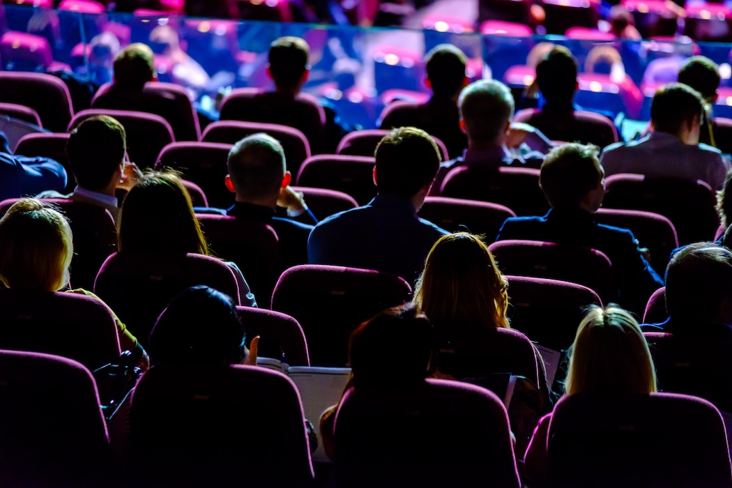 Audience members seated in a dimly lit conference hall with rows of red chairs, attentively watching a presentation or speaker on stage, illuminated by colorful stage lighting