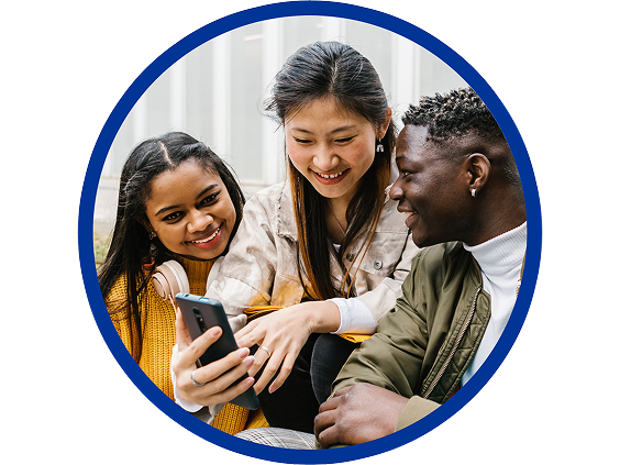 Three smiling students sitting together, looking at a smartphone screen, with one person holding the phone and the others gathered around, enjoying the moment