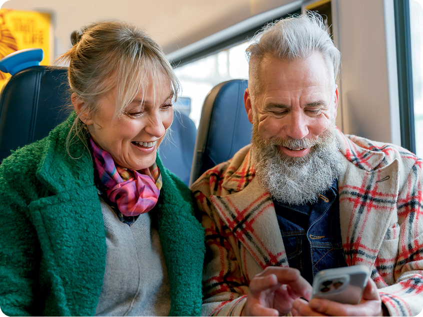 Smiling older couple sitting together on a train, looking at a smartphone and laughing