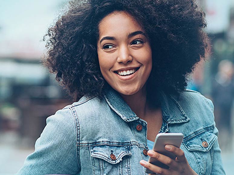 Smiling woman in a denim jacket sitting outside with a coffee cup in one hand and a phone in the other